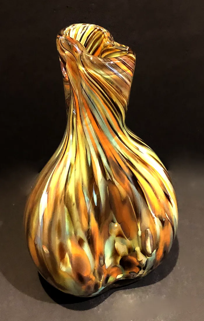 A blown glass vase in amber and gold with fine ribs spiralling up a folded, twisted body and dark speckles pooling at the base